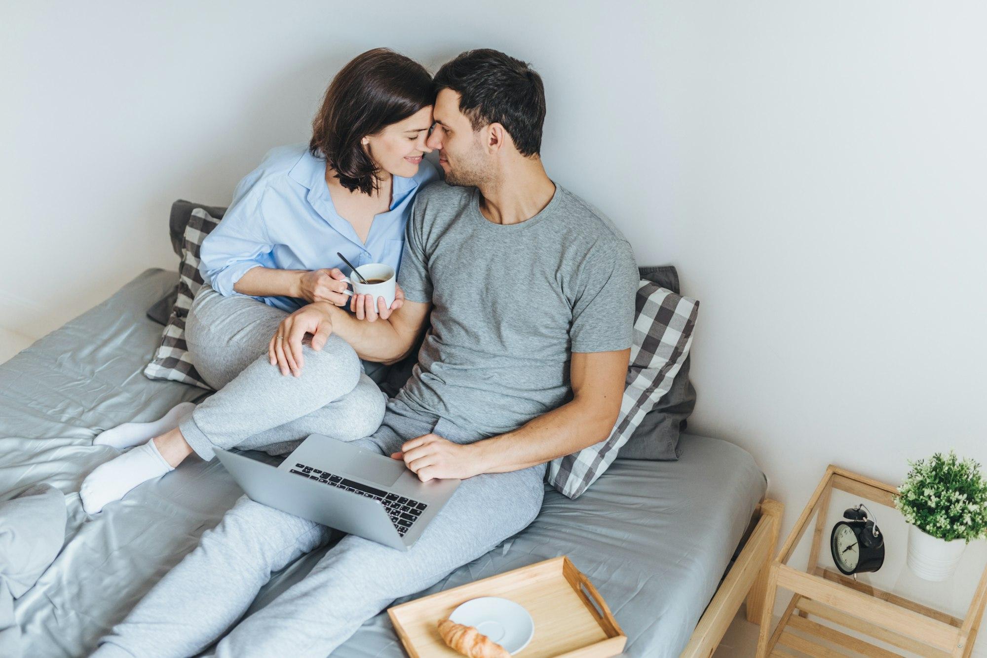 Couple going to kiss, look at each other as sit on bed, use laptop computer for watching films